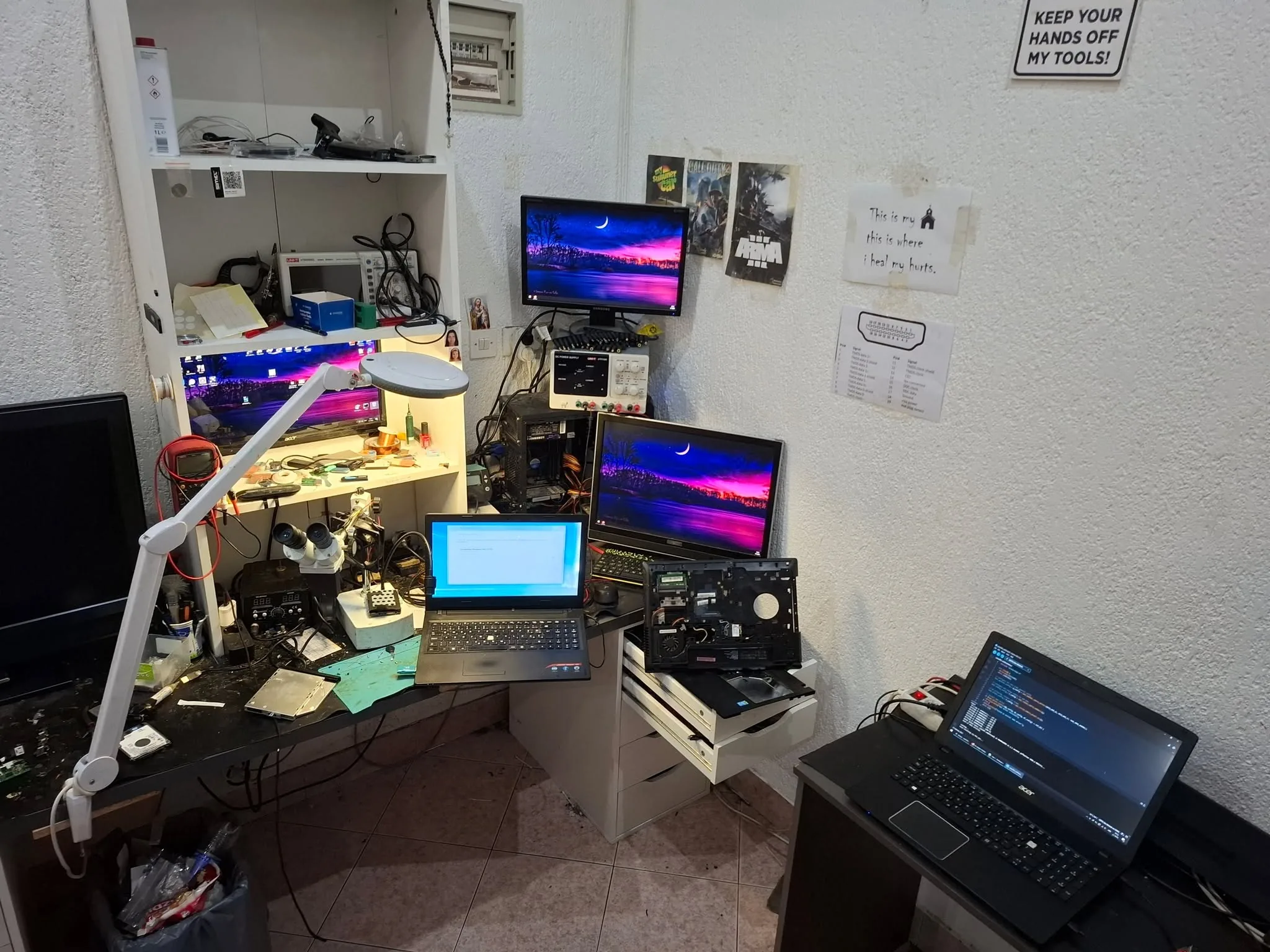The electronics repair bench — scopes, PSU, microscope, monitors and boards mid-repair